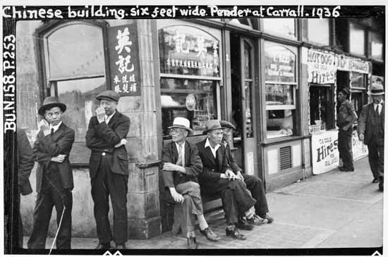 Black and white photo of a group of man standing and seated on the corner of a building