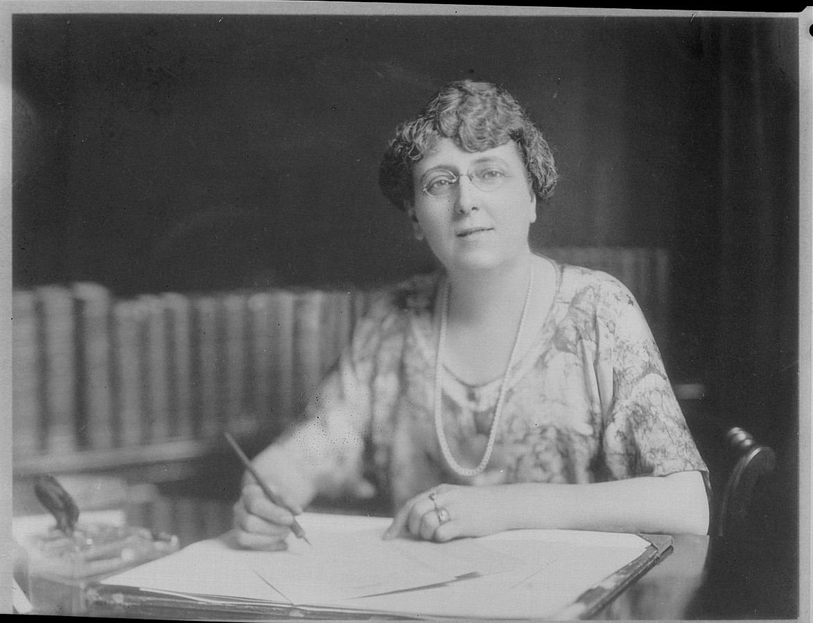 Black and white photo of a person seated at a desk and holding a pen hovering over an open book