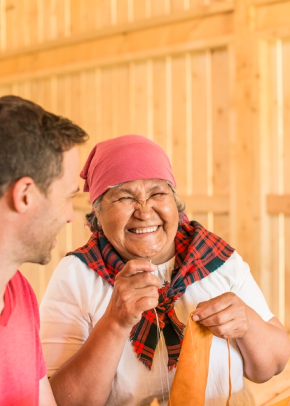 A person sewing fabric smiles and laughs while looking at another person who smiles back.