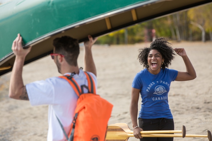 One person carries a canoe over their head while another person holding two paddles cheers them on.