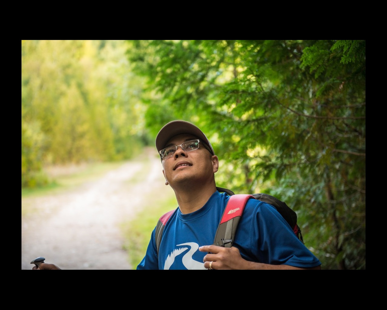 A close-up of a person smiling stands while looking upward on a forested trail.