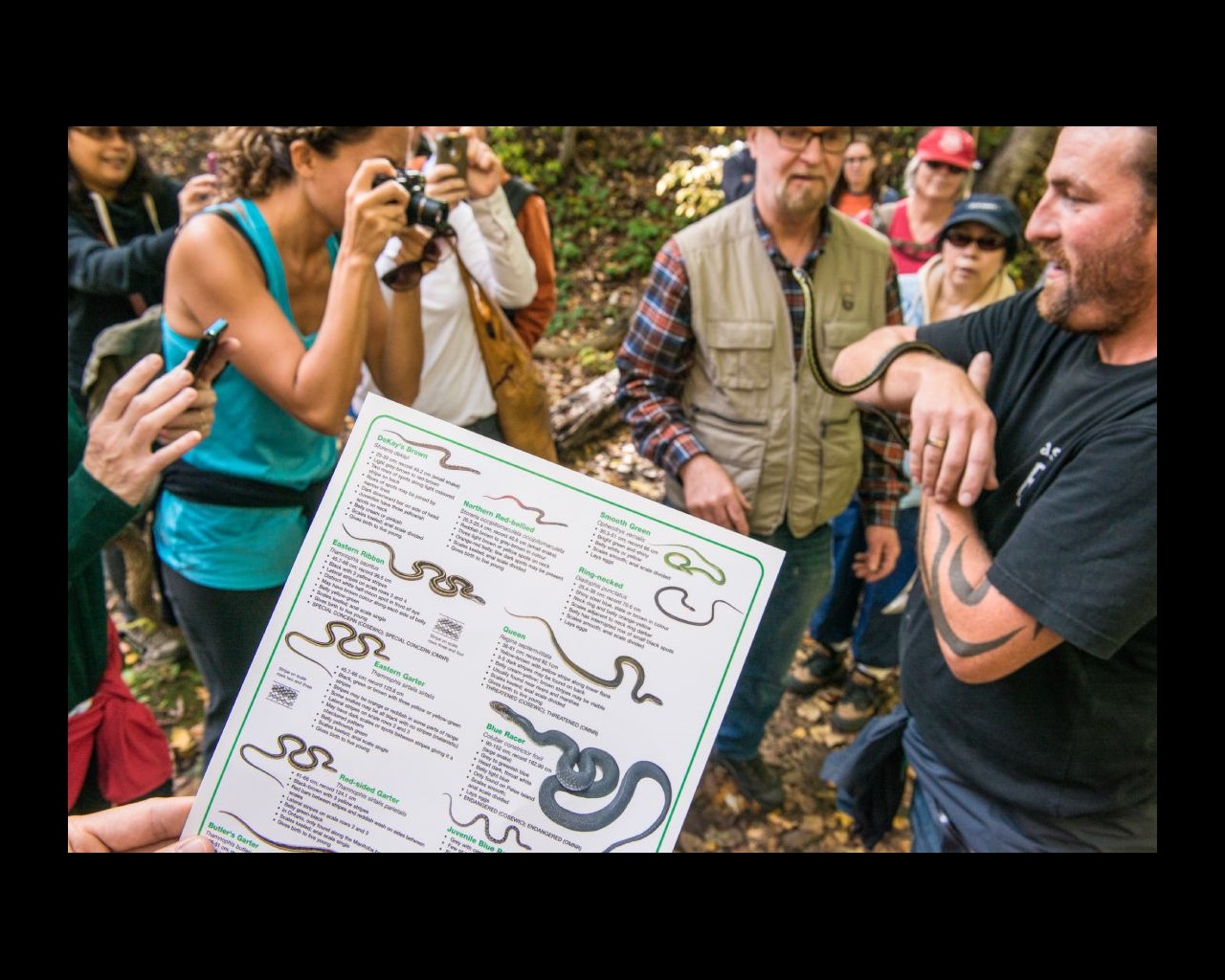 A group of people gather around a person holding a snake. One person holds a pamphlet of different snakes.