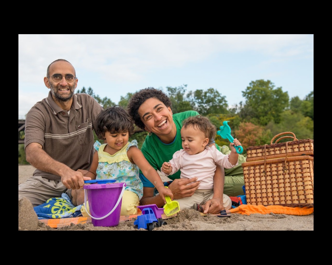 A family with two young children sit smiling on a picnic blanket while the children play in the sand.