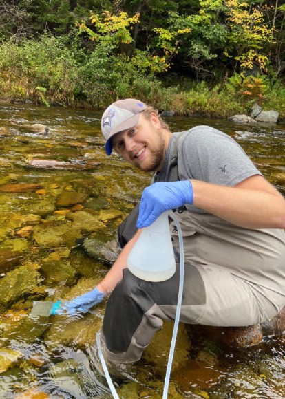 A researcher kneels in the water holding a plastic beaker and tube with gloved hands as they collect a water sample.