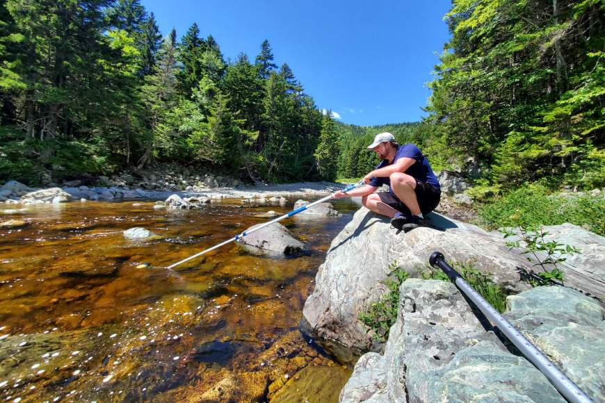 A researcher sits on a boulder at the edge of a river as they use a long pole in the water to collect a water sample.