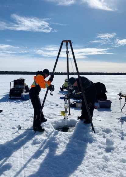 Two people in winter clothes lower a coring device attached to a large tripod into a hole cut in the thick snow-covered ice.