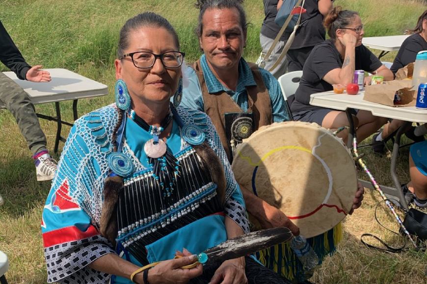 An Indigenous woman in ceremonial clothing sits next to her Indigenous partner who is holding a hand drum.