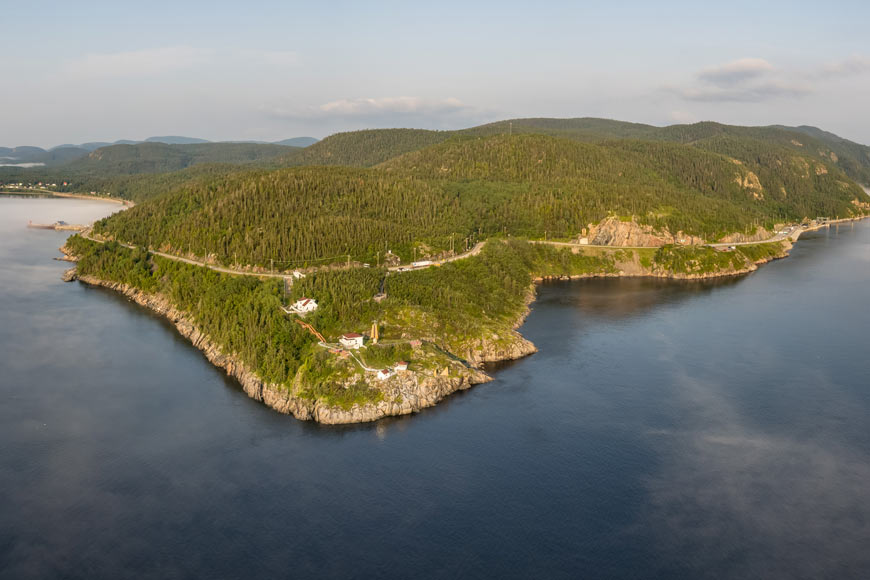 An aerial view of blue still water surrounding a tree-covered rolling peninsula that spans far into the distance.