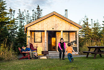 A child runs to his mother as two men sit in front of a rustic wooden cabin.