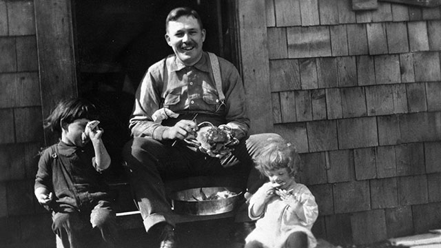 A man prepares crabs on the porch of a house with two young children by his side. 