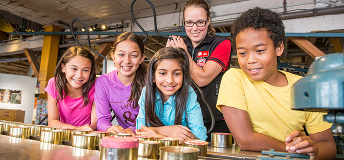 A group of children watch a presentation about a historical fish canning machine. 