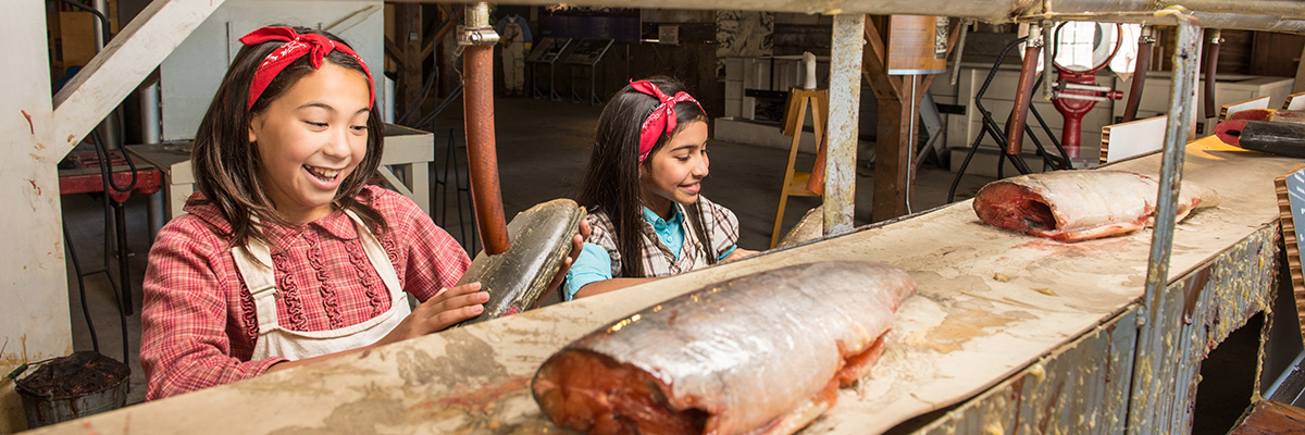 Two girls dressed up like original Cannery workers, pretending to be busy at work on the fish cleaning line.