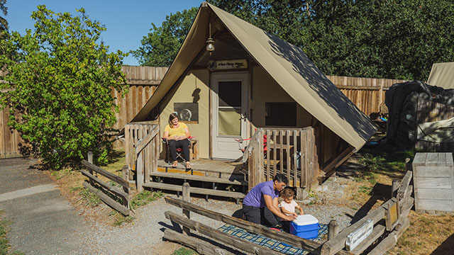 Wider shot of one parent sitting on a picnic blanket eating with child in front of an oTentik while the other parent watches them from a chair on the oTentik porch .