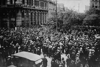 A black-and-white photo of a large group of people gathered on a street next to a tall building.