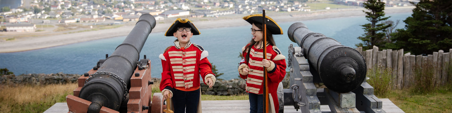 two children in historic military costume laughing next to two cannons overlooking a coastal town