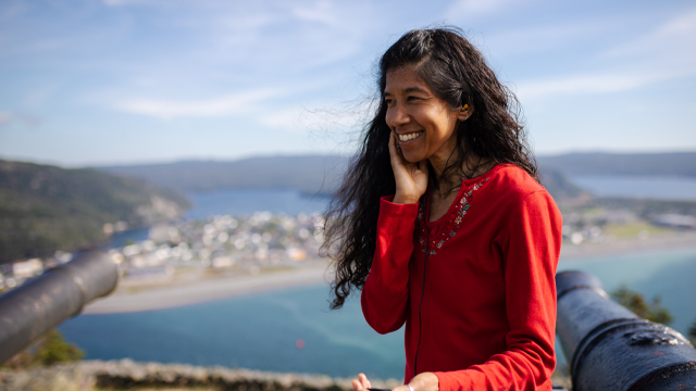 an individual in a red sweater, listens to an earphone while sitting overlooking an oceanside town