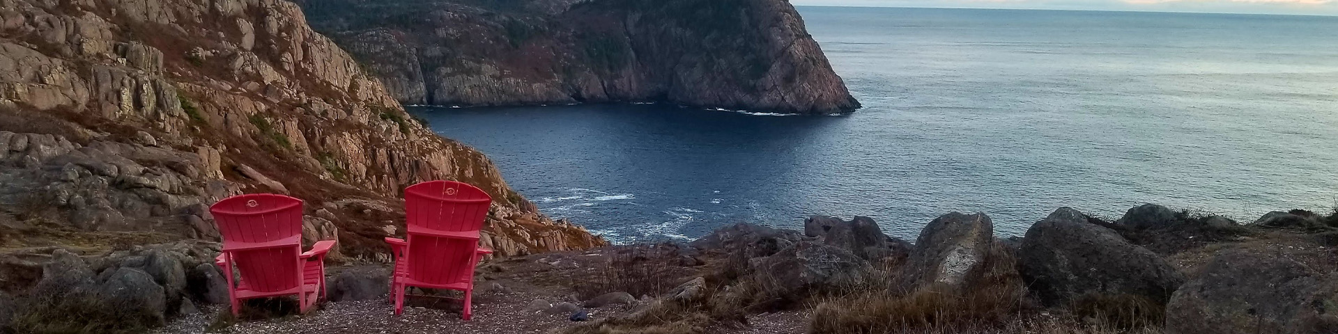 two red adirondack chairs, overlooking a rugged, ocean coastline