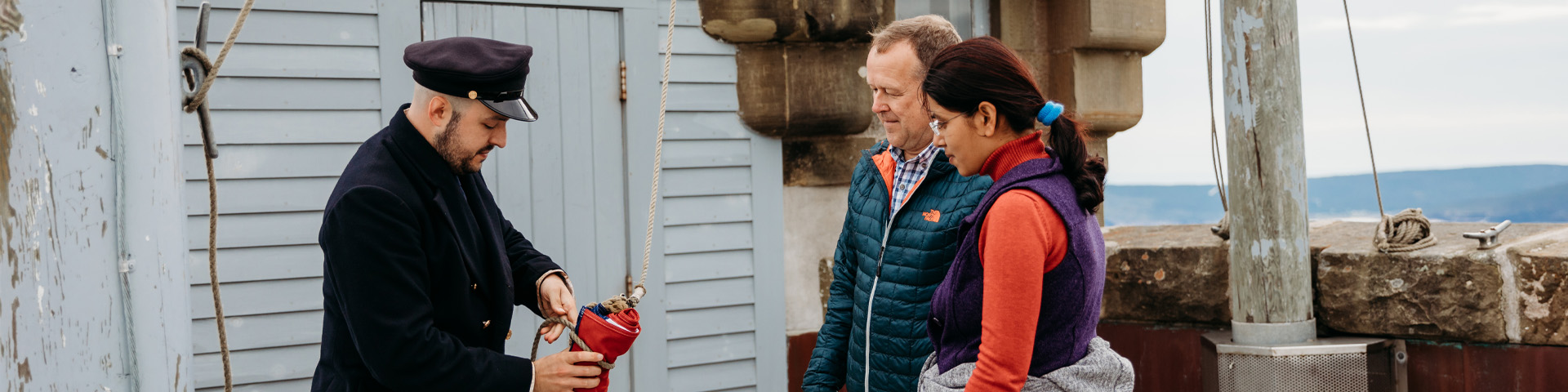 A Parks Canada employee in historic costume shows two individuals how to unfold a flag