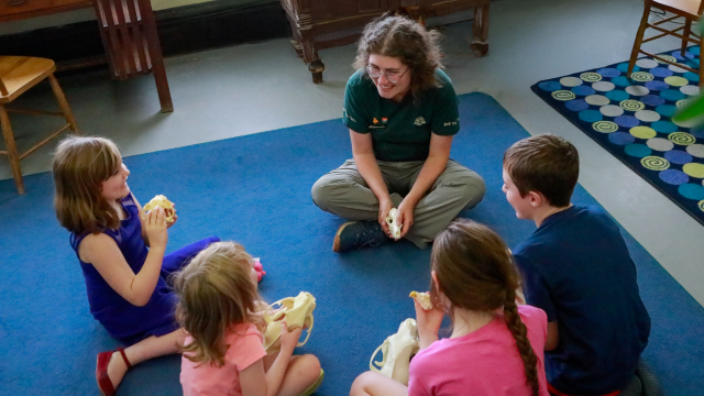 As part of an outreach program, students sit on the ground with a Parks Canada employee and hold objects in their hands.