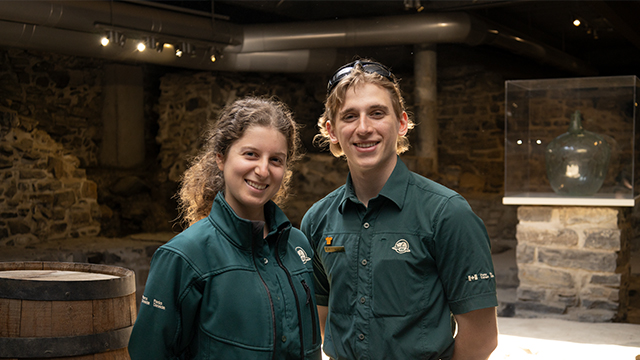 Two Parks Canada employees in uniform pose in front of the icebox at the -Saint-Louis Forts and Châteaux national historic site.