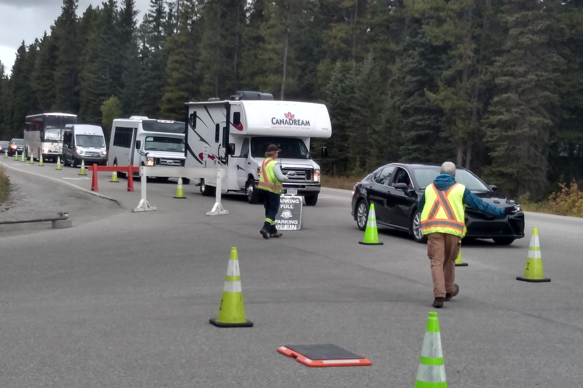 Two traffic flaggers redirect a lineup of traffic using their hands, pylons, and signage.  