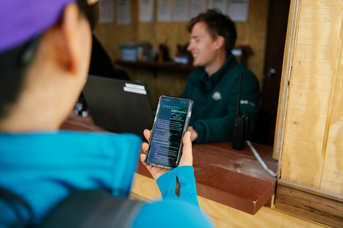 A visitor holds their phone up to a Parks Canada staff sitting in a wooden shelter. 