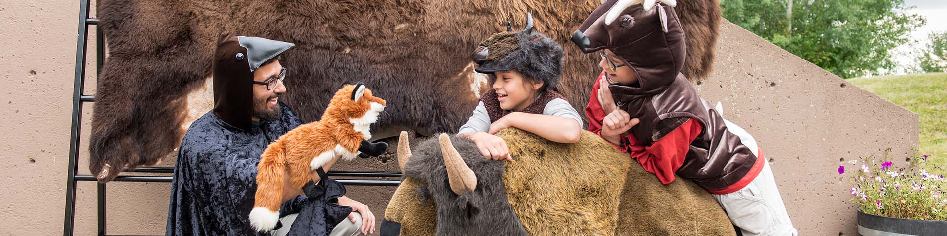 An interpreter in costume speaks to two children in costume in front of the Astotin Theatre, Elk Island National Park.