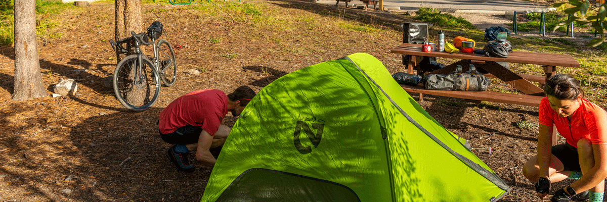 Two cyclists are pitching their tent. A bike and picnic table can be seen in the background.