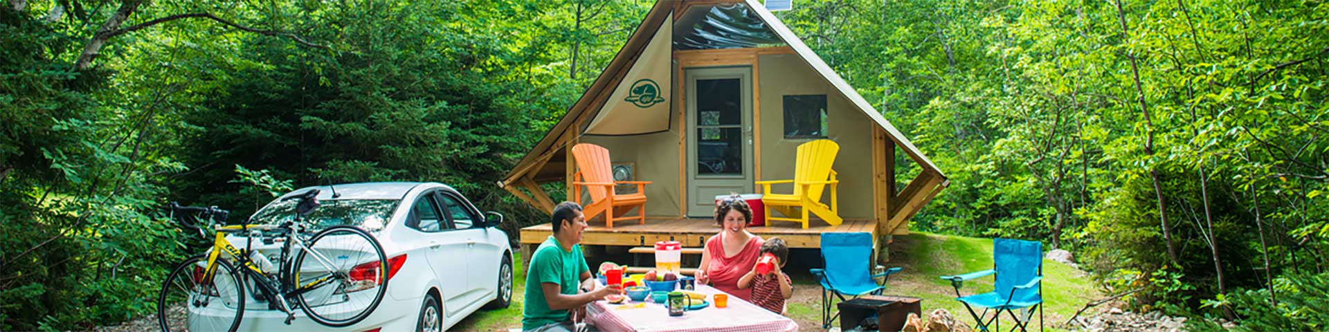 Deux adultes et un enfant sont assis à une table de pique-nique devant un Otentik de Parcs Canada, à gauche il y a une voiture garée avec un vélo à l'arrière et à droite un feu avec deux chaises.