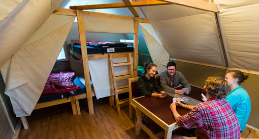 Four people playing cards at a table inside the oTENTik, with beds shown in the background.