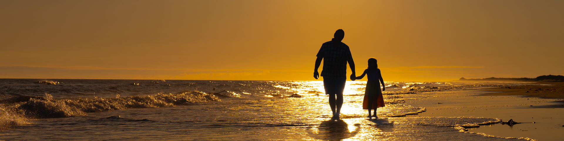 One and adult and one child walk hand in hand along the beach at sunrise.