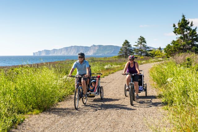 Two cyclists pulling children's trolleys ride along a gravel path with the cliffs in the distance.