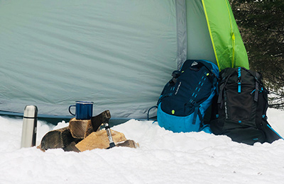 Backpacks, wood logs, an axe, a cup and a thermos lie in the snow in front of a tent.