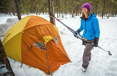 A person is carrying cross-country skis next to a tent set up in the snow, in a forest.