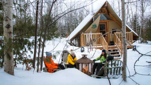 Des personnes sont assises autour d'un foyer extérieur dans la neige, devant un hébergement oTENTik.