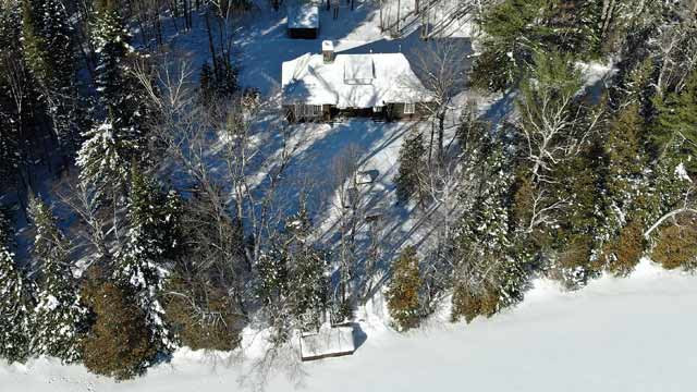 An aerial view of a Domaine Wabenaki-Andrew lodge next to a dock, by a lake, in a snow-covered forest.