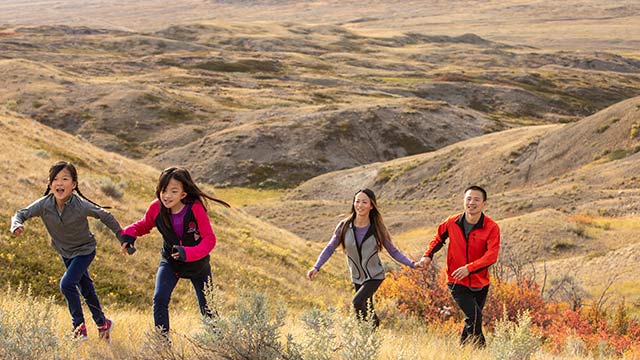 Two children running up a hill, with their parents following, with the landscape of the West Block of Grasslands National Park in the backround.