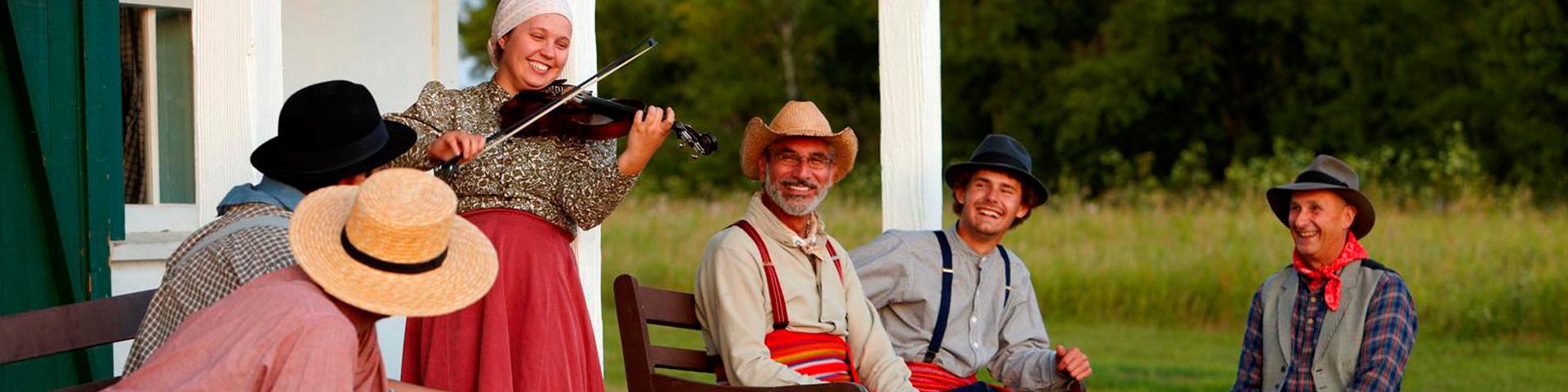 A costumed interpreter playing the violin on a terrace among other interpreters at Batoche National Historic Site.