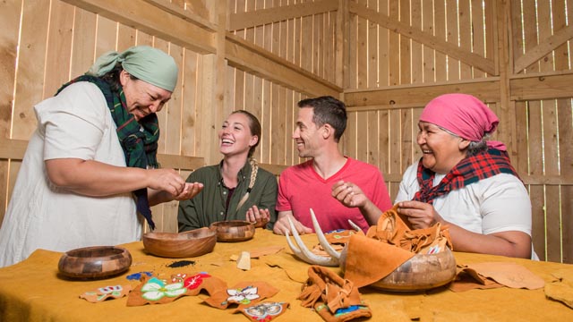 Two Indigenous women teaching beading and leather crafts to two young adults inside the Boat House at Rocky Mountain House National Historic Site.