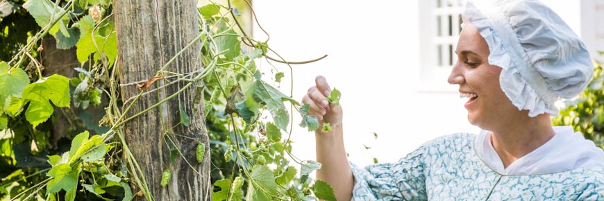 Une guide de Parcs Canada en costume d'époque s'occupe du jardin, le sourire aux lèvres, par une journée ensoleillée.