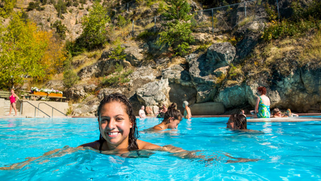 A young woman soaking in the Radium Hot Springs with visitors and a cliff in the background