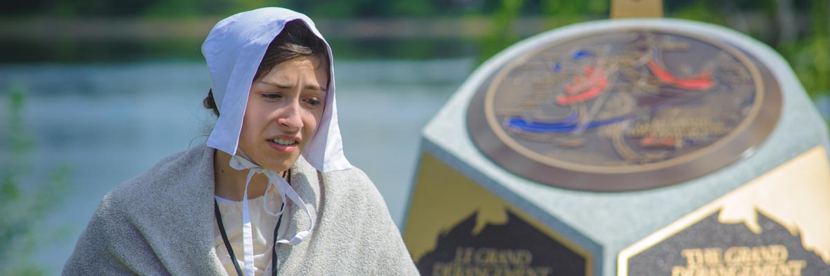 Une femme en costume d’époque devant un monument sur le Grand Dérangement.