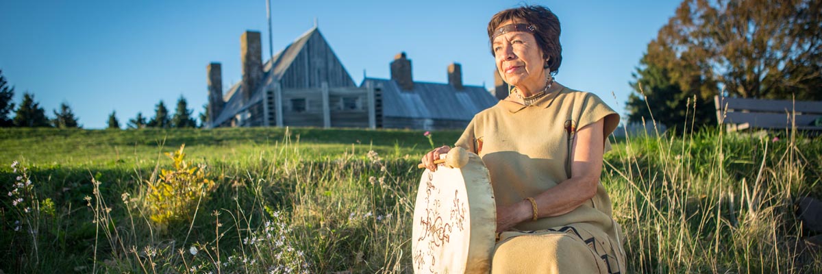 Une femme autochtone vêtue d’un costume mi’kmaw traditionnel est assise avec un tambour parmi les rochers à l’extérieur de l’Habitation.